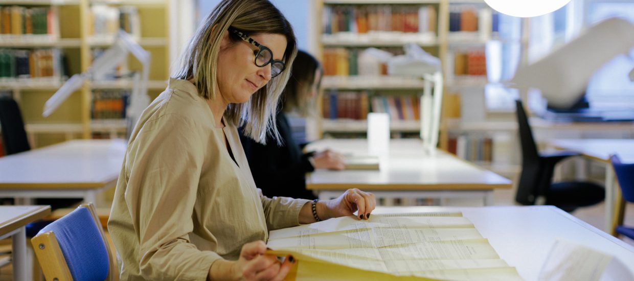 Kvinne med langt hår og briller sitter ved en pult på en lesesal og ser på et stort ark med skrift. Pulter og bokhyller i bakgrunnen