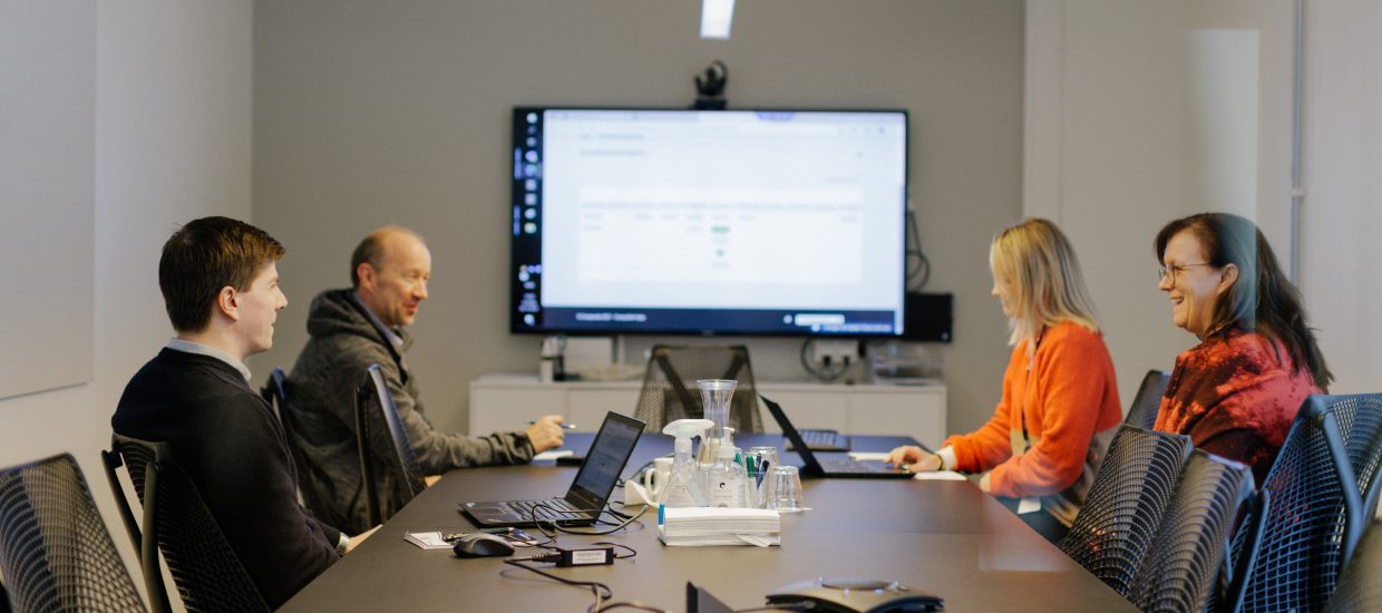 To menn og to kvinner sitter i ved et bord i et møterom. Datautstyr på bordet og skjerm med uklart innhold på veggen.