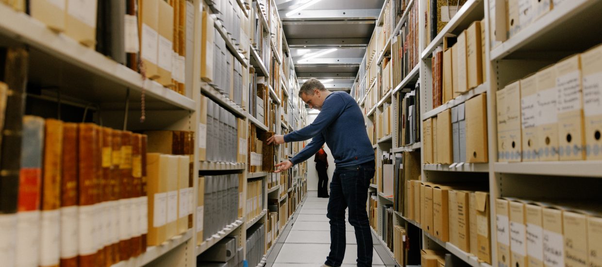 Mann står mellom to arkivreoler med arkivbokser og protokoller.