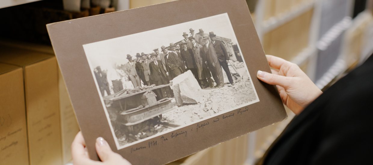 Hender holder svart-hvitt bilde av en gruppe menn. Arkivbokser på hylle i bakgrunnen.