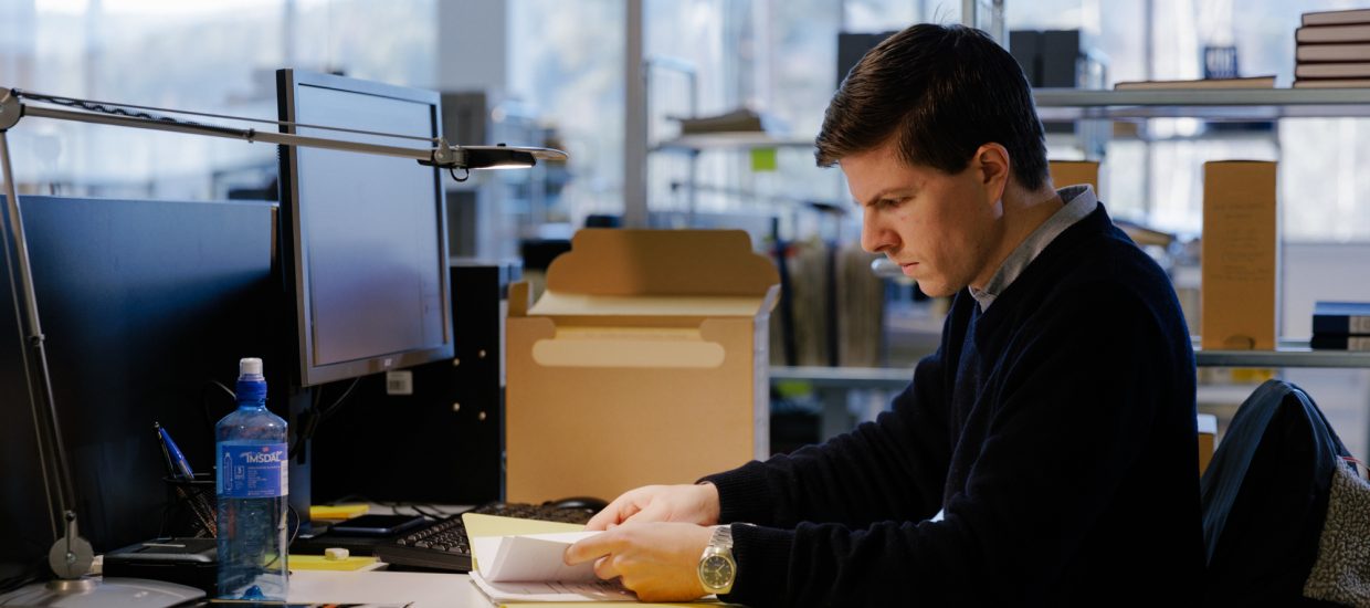 Mann med kort brunt hår og blå genser sitter ved en pult og blar i dokumenter. Arkivboks står på pulten.