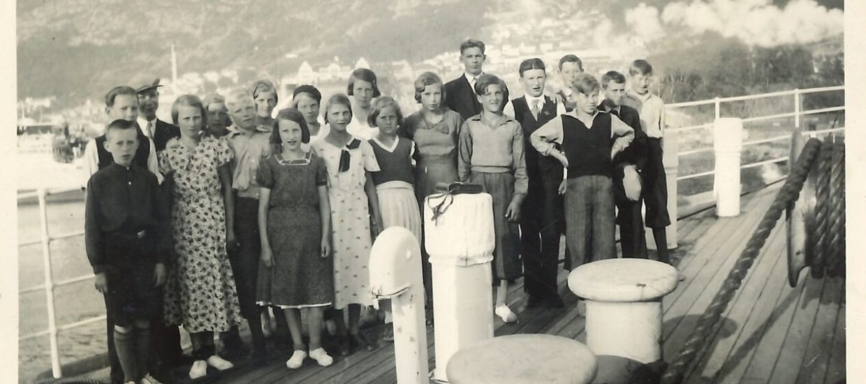 Unge gutter og jenter står oppstilt på en båt. Bebyggelse og fjell i bakgrunnen.