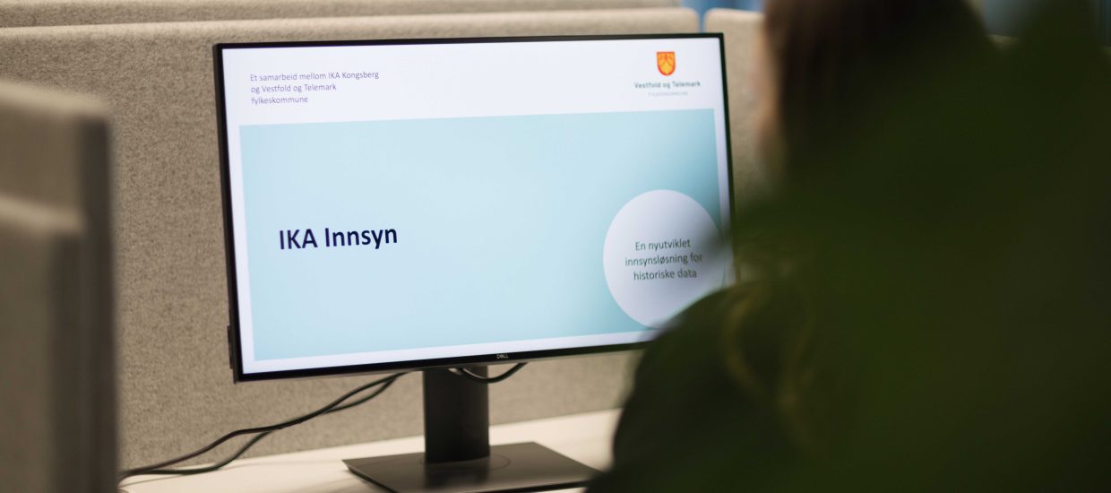 En pc-skjerm med teksten «IKA Innsyn» og en ansatt som sitter og jobber med et skrivebord i et kontorlandskap.