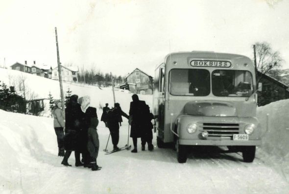 Bokbussen på snødekt vei med høye brøytekanter, barn og voksne ved bussen. En person på ski. 1952