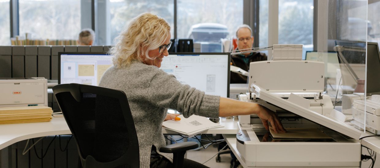 en dame med blondt, krøllete hår og grå ullgenser smiler og tar ut et ark fra en kopimaskin i et kontorlandskap