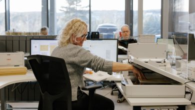 en dame med blondt, krøllete hår og grå ullgenser smiler og tar ut et ark fra en skanner i et kontorlandskap