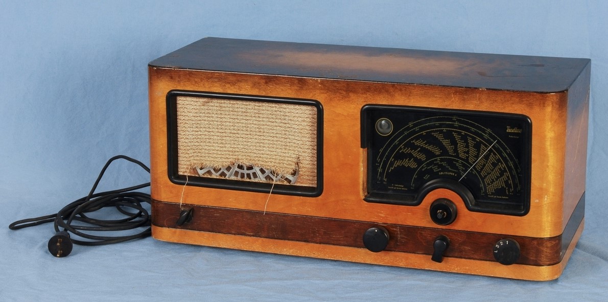 Radio fra 1947 i brunt tremateriale, svarte knotter og frekvensviser. Slitt stoff foran høyttaleren. 