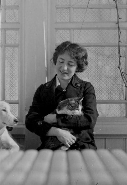 Kvinne sitter og holder en katt i armene, og en hund ved siden av seg. svart hvitt bilde.