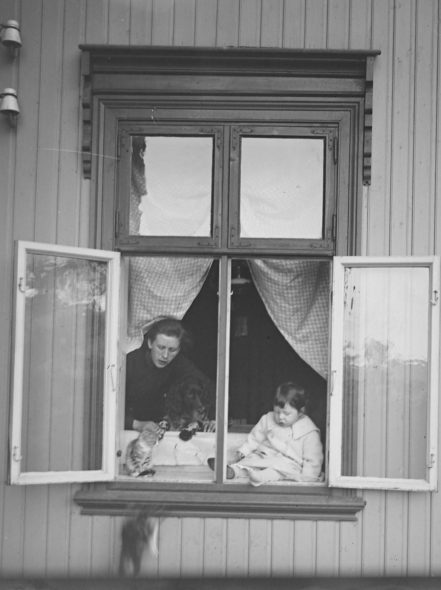 En kvinne, et barn og en katt i et vindu. Barnet sitter i vinduskarmen, og en katt faller ut av vinduet. Svart hvitt bilde.