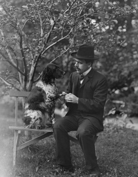 Mann med dress og hatt sitter på benk i hage med en hund ved siden av seg. Han holder hunden i poten. Svart hvitt bilde