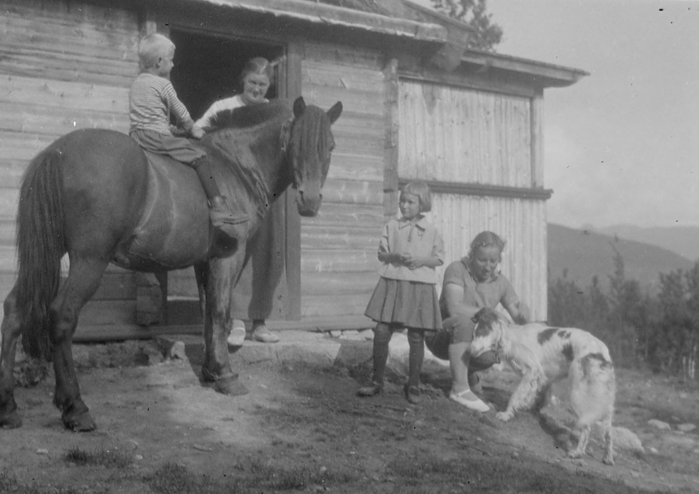 To kvinner, en gutt og en jente utenfor bygning. Gutten sitter på hest, og den ene kvinnene klapper en hund. svart hvitt bilde.