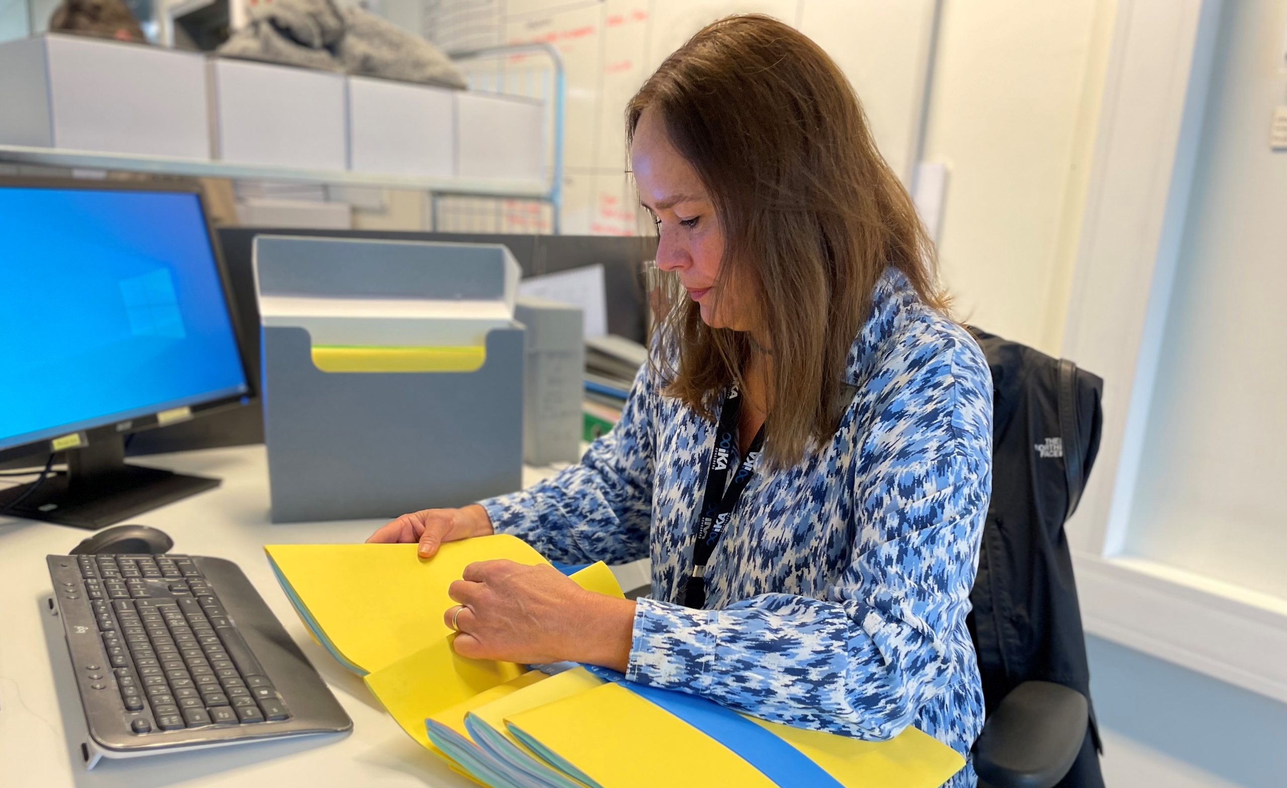 Kvinne med langt, brunt hår og blå og hvit kjole sitter ved en pult. Hun ser på pulten, og har gule mapper i hendene med et blått alfabetiseringsverktøy.